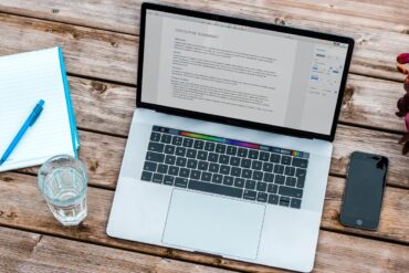 Silver gray macbook beside spacegray iPhone 6 and clear drinking glass on brown wooden top